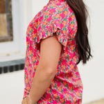 Woman with dark hair standing in a side profile position wearing the Stevie Top against a white background. The focal point of this image is the detailing in pink and orange floral as well as the ruffles on sleeves. The model paired this top with dark-wash denim jeans. 