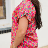 Woman with dark hair standing in a side profile position wearing the Stevie Top against a white background. The focal point of this image is the detailing in pink and orange floral as well as the ruffles on sleeves. The model paired this top with dark-wash denim jeans. 