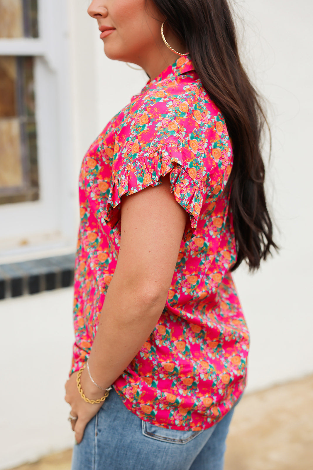 Woman with dark hair standing in a side profile position wearing the Stevie Top against a white background. The focal point of this image is the detailing in pink and orange floral as well as the ruffles on sleeves. The model paired this top with dark-wash denim jeans. 