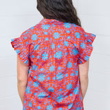 Woman with dark hair standing in a back profile position wearing the Stevie Top against a white background. The focal point for this image is the pleated back and graphics. The model paired this top with dark-wash denim jeans. 