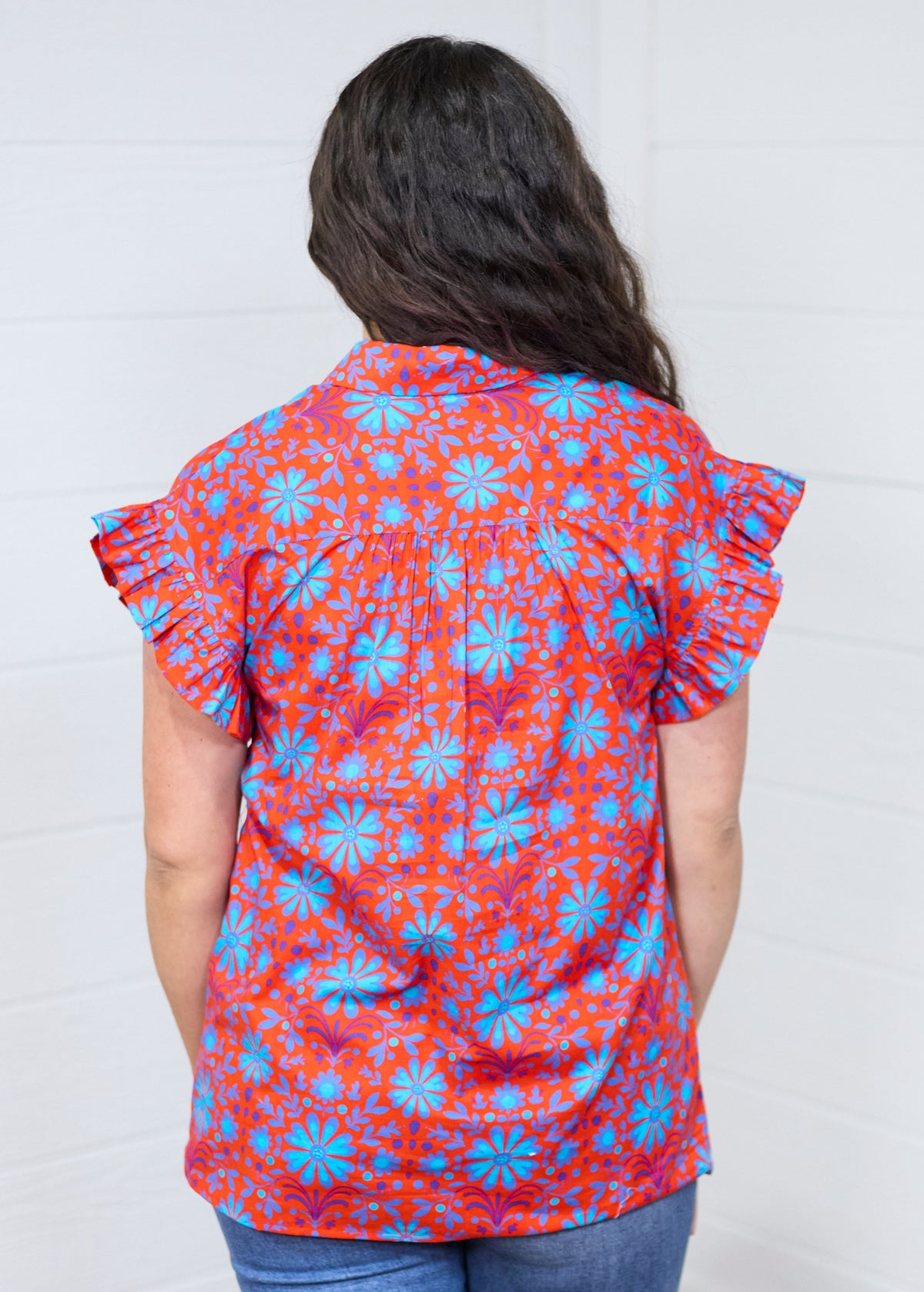 Woman with dark hair standing in a back profile position wearing the Stevie Top against a white background. The focal point for this image is the pleated back and graphics. The model paired this top with dark-wash denim jeans. 