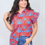 Woman with dark hair standing in a front profile position wearing the Stevie Top against a white background. The focal point of this image is the graphics on top, ruffled sleeve and buttons down front of shirt. The model paired this top with dark-wash denim jeans. 