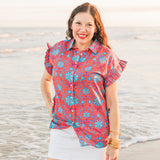 Woman with dark hair standing in a front profile position wearing the Stevie Top in a lifestyle scene. The focal point of this image is the shirt in its entirety; buttons, ruffles, graphics, low collar. The model paired this top with a white skirt.