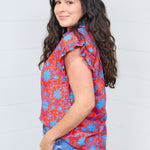 Woman with dark hair standing in a side profile position wearing the Stevie Top against a white background. The focal point of this image is the ruffle cuff and red/blue floral graphics. The model paired this top dark-wash denim jeans. 
