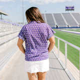 Woman with dark hair standing in back profile wearing the Stevie Top at a football field. The focal point for this image is the graphics on the back of the top as well as the striped ruffle on sleeves. The Model paired this top with a white denim skirt.