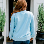 Woman with red hair wearing a baby blue sweatshirt with Christmas motif embroidery. Model paired this top with dark-wash denim jeans. 