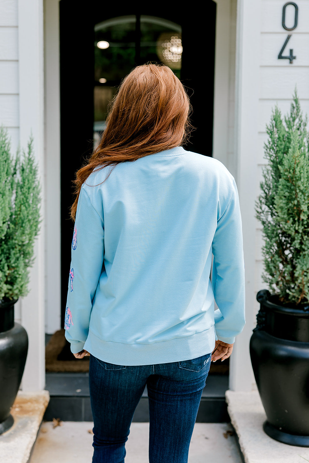 Woman with red hair wearing a baby blue sweatshirt with Christmas motif embroidery. Model paired this top with dark-wash denim jeans. 