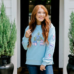 Woman with red hair wearing a baby blue sweatshirt with Christmas motif embroidery. Model paired this top with dark-wash denim jeans. 