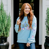 Woman with red hair wearing a baby blue sweatshirt with Christmas motif embroidery. Model paired this top with dark-wash denim jeans. 