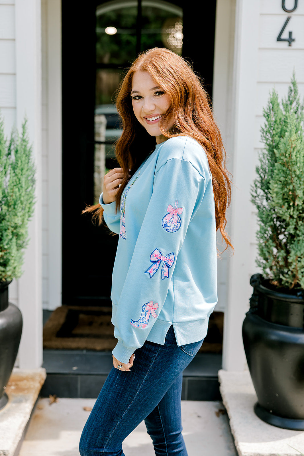 Woman with red hair wearing a baby blue sweatshirt with Christmas motif embroidery. Model paired this top with dark-wash denim jeans. 