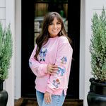 Woman with dark hair wearing a baby pink sweatshirt with embroidered Christmas motifs on front and sleeves of top. Model paired this top with light-wash denim jeans. 