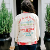 Woman with dark hair wearing a sweatshirt with red & white striping along cuff and collar. On the back of top there is a multi color bible verse. Model paired this top with dark-wash denim jeans. 