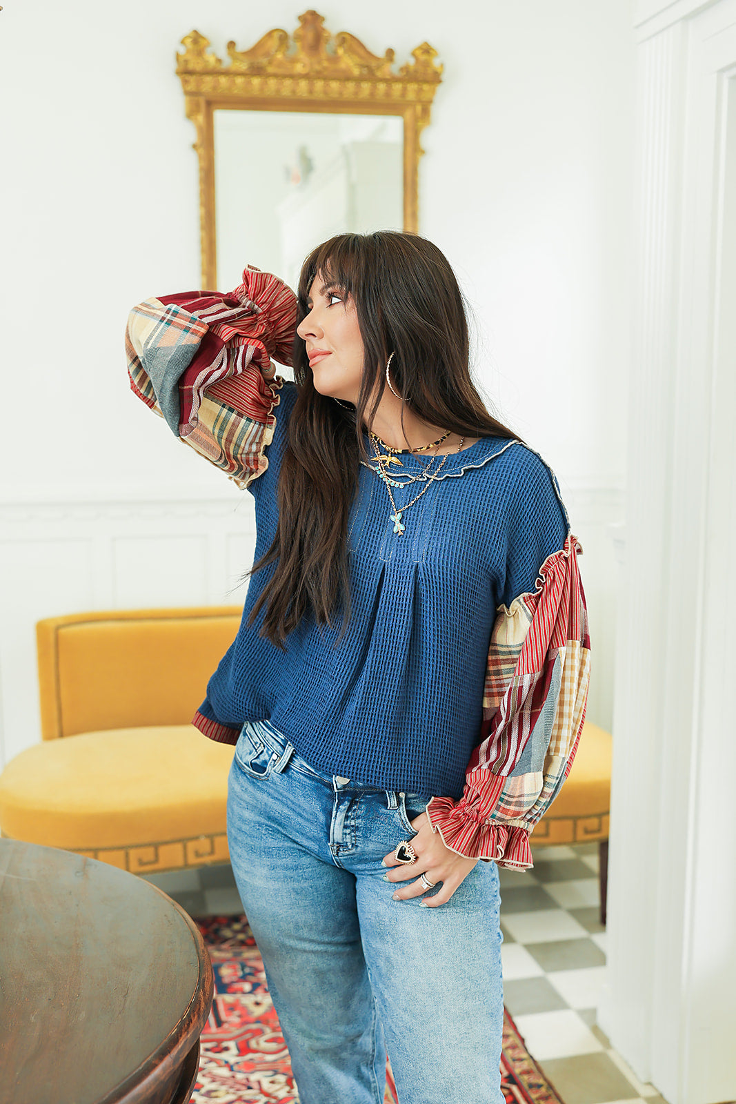Woman with dark hair standing in a front profile position wearing the Tammy Top in a lifestyle scene. The focal point of this image is the waffle knit fabric and puffy sleeves. The model paired this top with mid-wash denim jeans. 