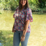 Woman with short dark hair standing in a front profile position wearing the Mia Top in a lifestyle scene. The focal point of this image is the Mia Top in  a heavy greenery scene. The model paired this top with light-wash denim jeans.