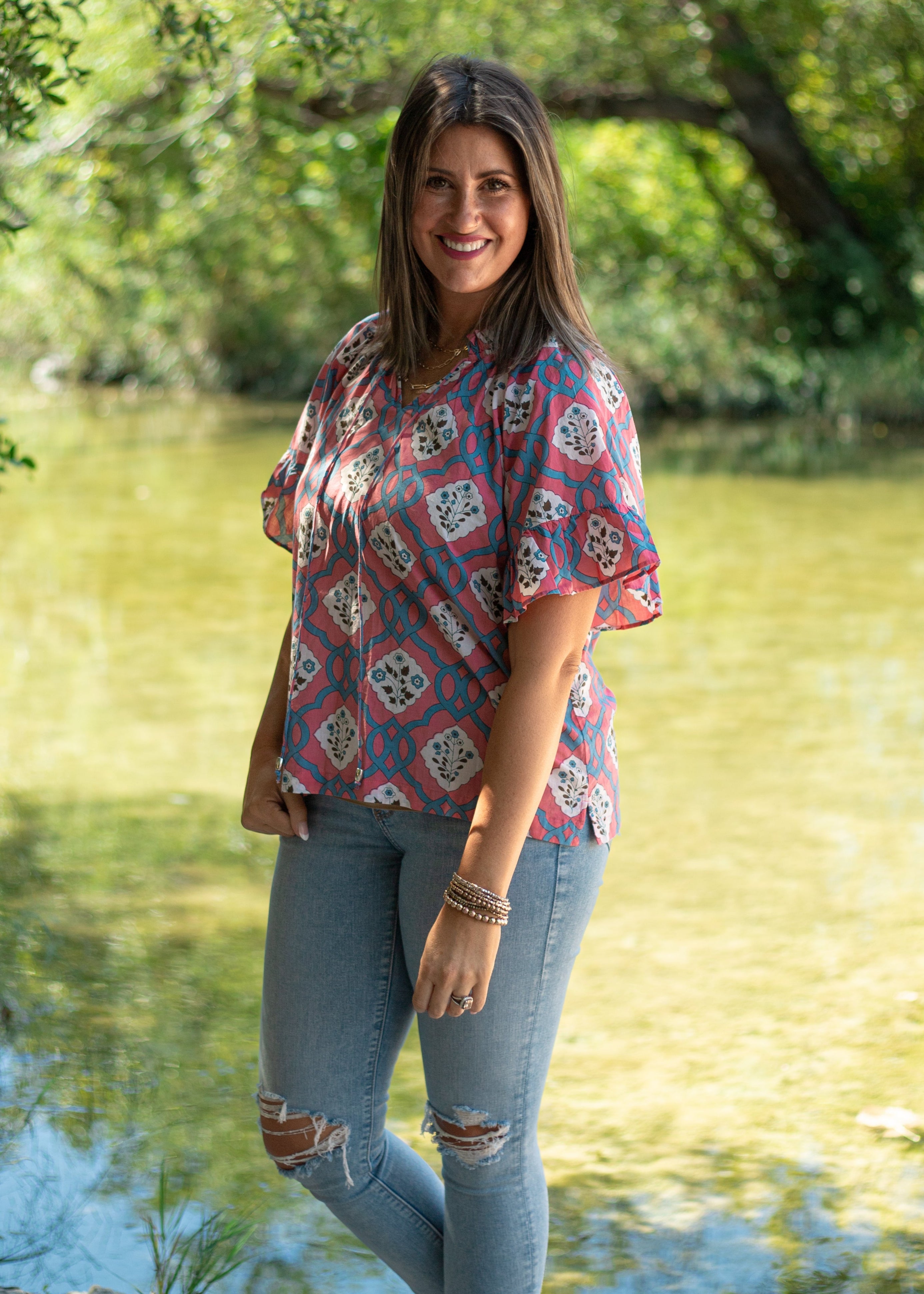 Woman with short dark hair standing in a front profile position wearing the Mia Top in a lifestyle scene. The focal point of this image is the Mia Top in  a heavy greenery scene. The model paired this top with light-wash denim jeans.