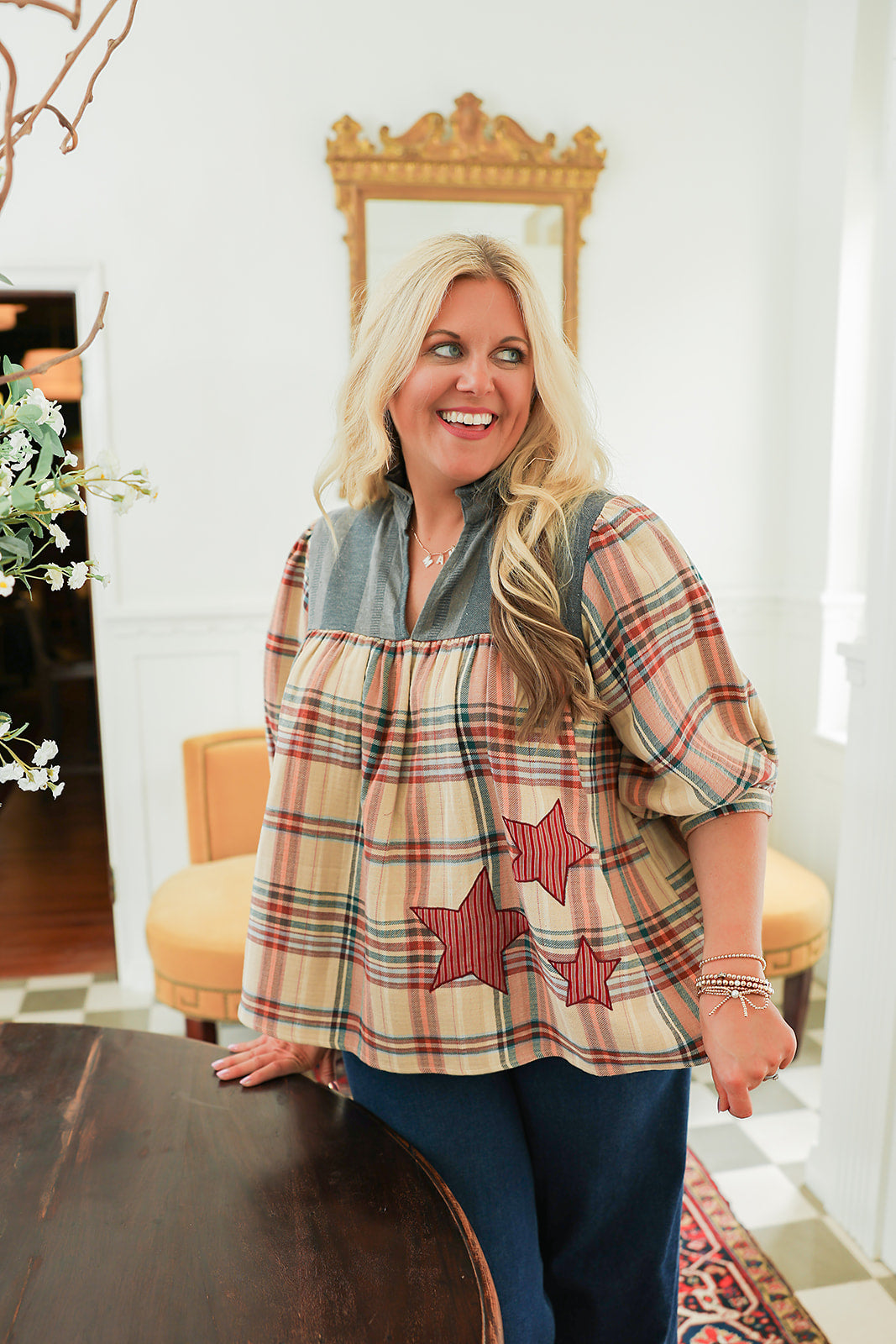 Woman with blonde hair wearing a quarter sleeve top with plaid patch-work paired with dark-wash denim jeans. 