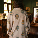 Woman with dark hair wearing a quarter sleeve poncho styled top that showcases yellow and blue floral graphics. Model paired this top with light-wash denim jeans. 