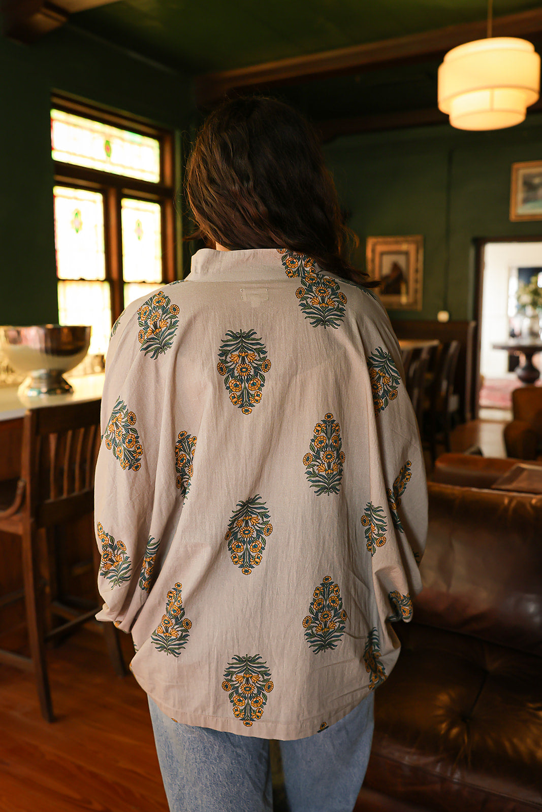 Woman with dark hair wearing a quarter sleeve poncho styled top that showcases yellow and blue floral graphics. Model paired this top with light-wash denim jeans. 