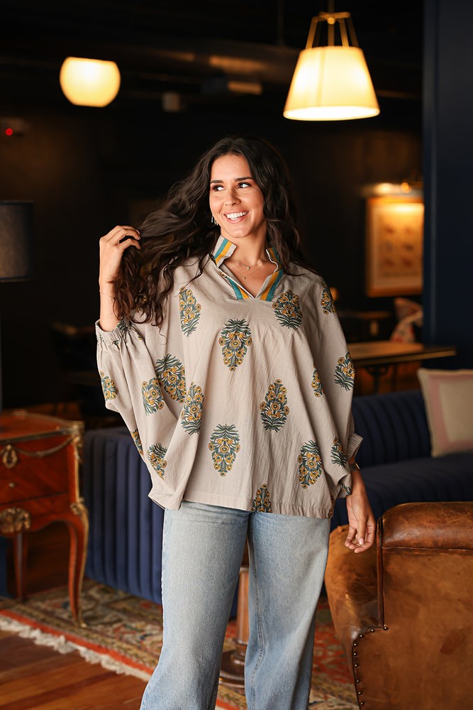 Woman with dark hair wearing a quarter sleeve poncho styled top that showcases yellow and blue floral graphics. Model paired this top with light-wash denim jeans. 