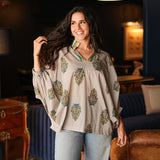 Woman with dark hair wearing a quarter sleeve poncho styled top that showcases yellow and blue floral graphics. Model paired this top with light-wash denim jeans. 
