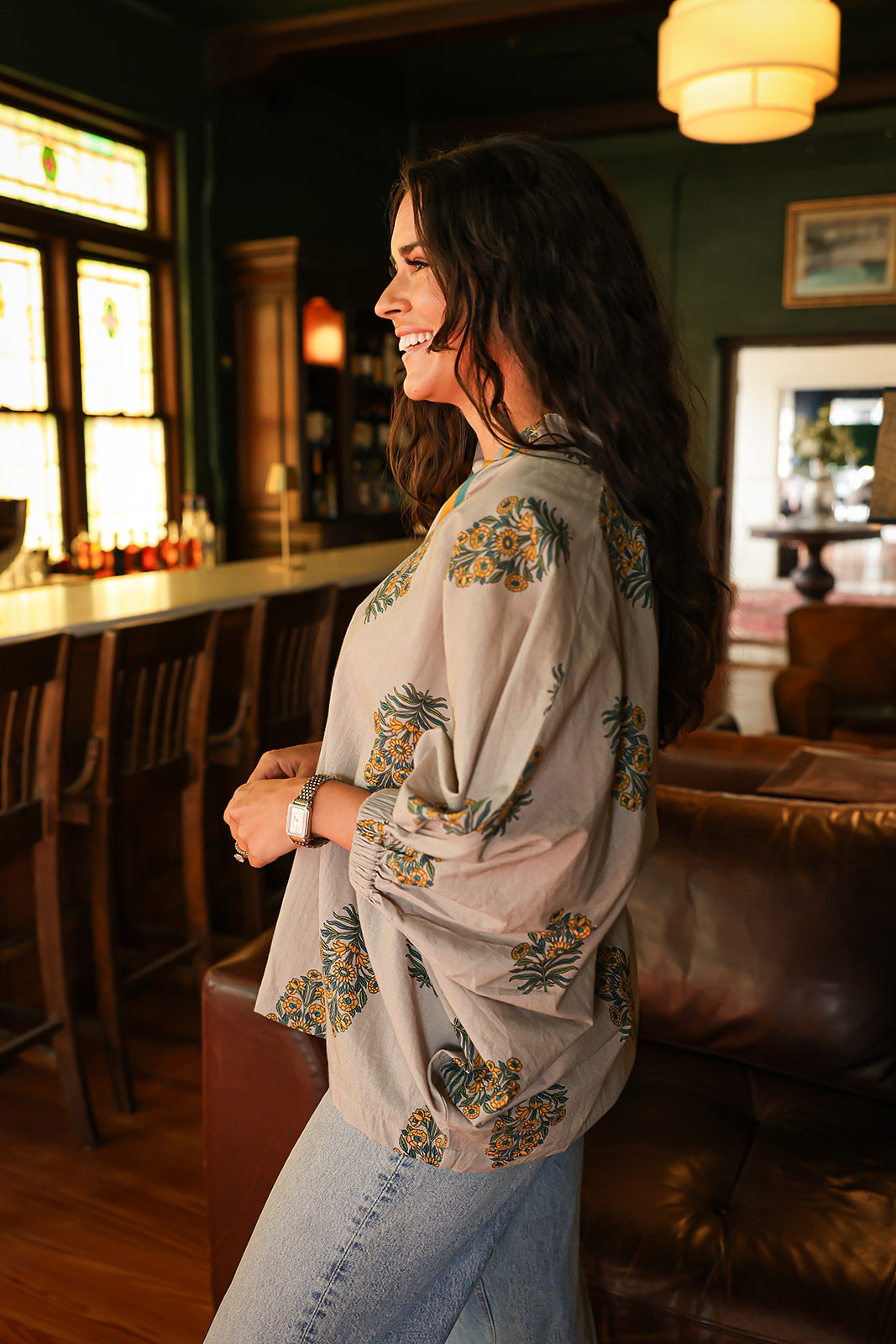 Woman with dark hair wearing a quarter sleeve poncho styled top that showcases yellow and blue floral graphics. Model paired this top with light-wash denim jeans. 