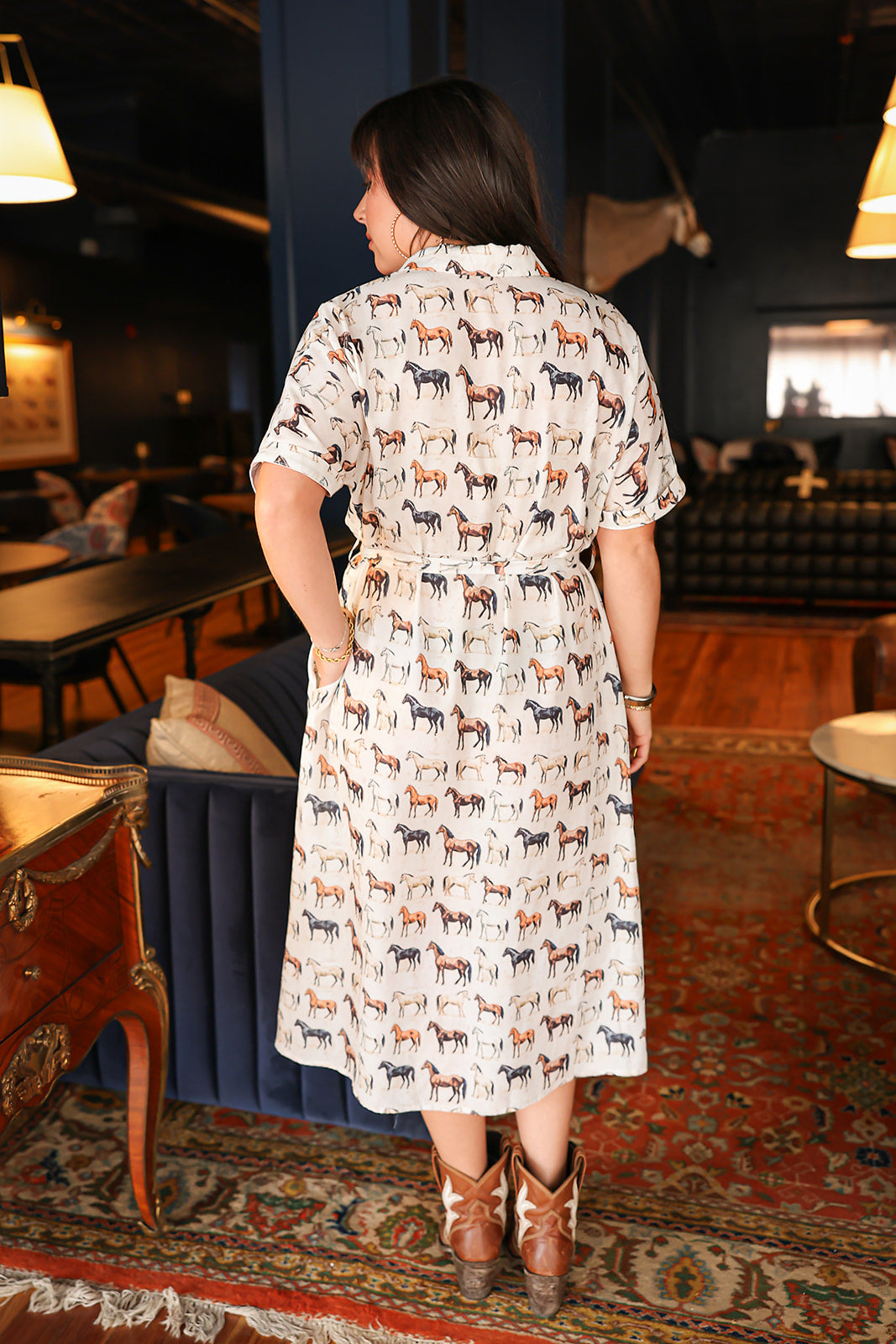 Woman with dark hair wearing a short-sleeve midi dress with multi-colored horses and belt to tie around wait. 