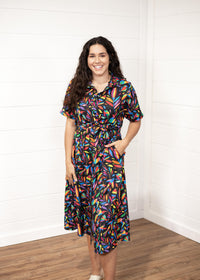 Woman with dark hair in front profile position wearing the Vanessa Dress against a white background. The focal point of this image is the front graphics on the dress with emphasis on the collar and belt. Model paired white sandals with this dress.