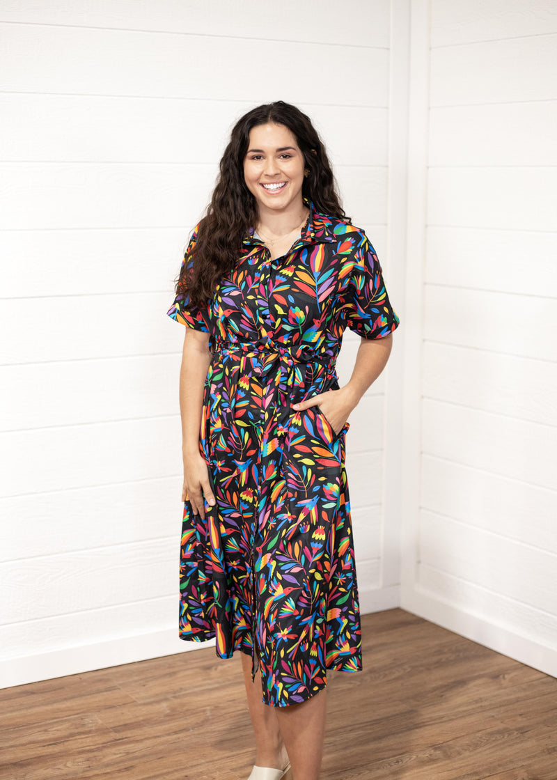 Woman with dark hair in front profile position wearing the Vanessa Dress against a white background. The focal point of this image is the front graphics on the dress with emphasis on the collar and belt. Model paired white sandals with this dress.