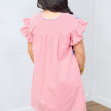 Woman with dark hair standing in a back profile position wearing the Wren Dress against a white background. The focal point of this image is the red gingham patter as well as the blue embroidery around neckline. 