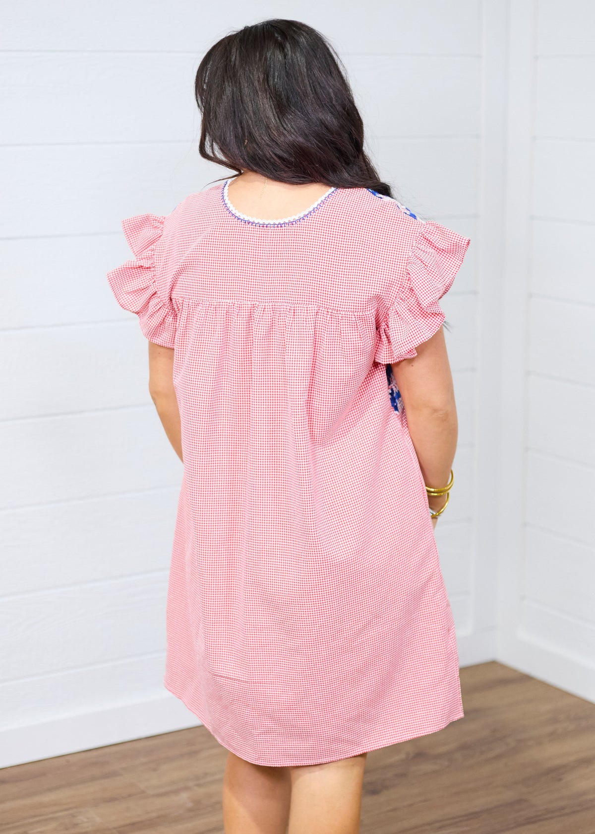 Woman with dark hair standing in a back profile position wearing the Wren Dress against a white background. The focal point of this image is the red gingham patter as well as the blue embroidery around neckline. 