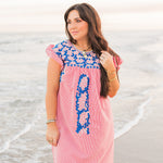 Woman with dark hair standing in a front profile position wearing the Wren Dress in a lifestyle scene. The focal point of this image is the dress in its entirety; gingham pattern, embroidery, and ruffle sleeves. 