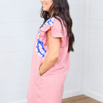 Woman with dark hair standing in a side profile position wearing the Wren Dress against a white background. The focal point of this image is the side of dress graphics, blue and white embroidery as well as ruffle sleeves. 