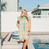 Woman with blonde hair standing in a front profile position wearing the Wren Dress against a "real time" background. The focal point of this image is to see the dress in its entirety as well as in real settings that add color and light. The model paired this dress with orange heels.